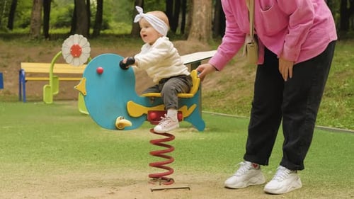 Mother Swings Her Child on Spring Riders on Playground in the Park Outdoors