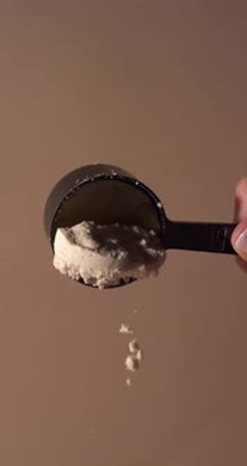Slow-motion close-up of or vanilla protein powder gently falling from a black scoop against a warm b