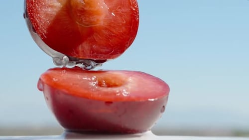Two halves of a red plum, water dripping on them, against a blue sky background. Close-up.