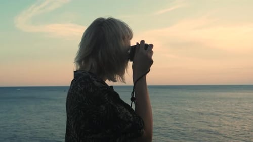 Young blonde woman takes pictures of the sunset sea with an analog camera.