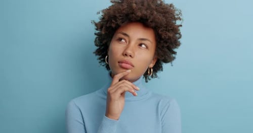 Serious Dark Skinned Woman with Thoughtful Expression Keeps Finger on Face Concentrated