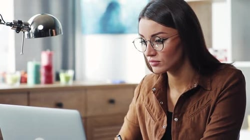 Attractive Smart Woman Sitting at Laptop Making Notes in Notebook Nice Girl in Glasses Finishing