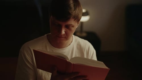 Portrait of Focused Young Man Enjoying Reading Interesting Book at Home Sitting Alone in Dark Room
