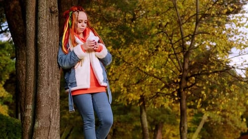 Young Adult Drinks Coffee Leaning on Tree in Autumn