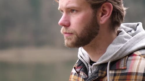 a close up of a face of a blond young man with a beard. he walks along a river which reflects the fo