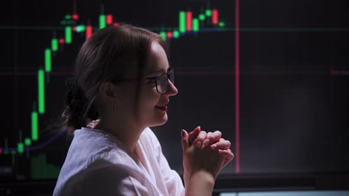 A Female Trader Speaks and Gestures with Her Hands Against Screen with Charts