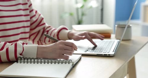 Young student studying with laptop and writing in notebook at wooden table indoors, closeup