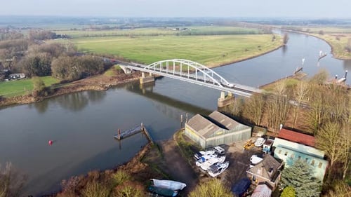 A white arched bridge spans a calm river curving through open fields, with boats, sheds, and small