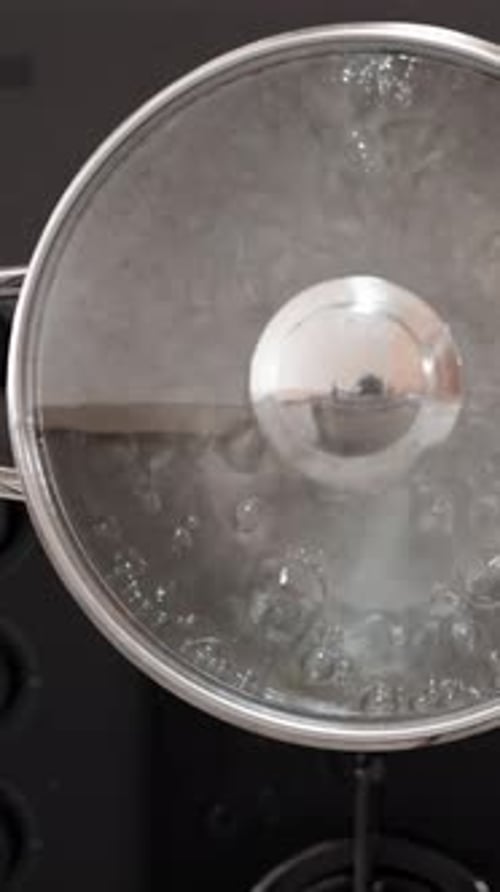 Vertical video. From a top view, I see a silver pot filled with boiling water on a black glass gas