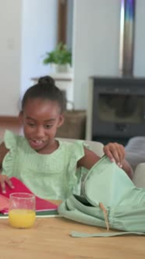 Vidéo verticale : soulevant un dossier rose, enfant en robe verte emballant un sac à dos menthe à table pour la classe