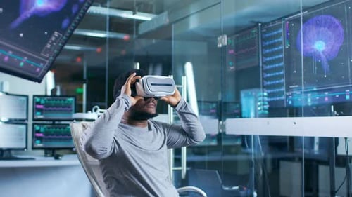 In Laboratory Scientist Wearing Virtual Reality Headset Sitting in a Chair Interacts With Monitors
