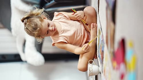 Little Girl Sits on the Floor at Home and Draws with Paints and Brushes in a Coloring Book