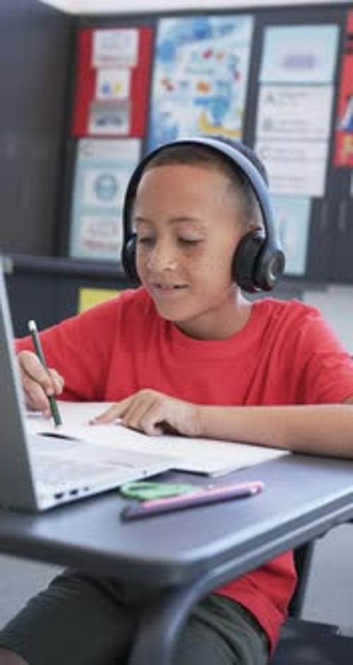 Vertical video: In school, child wearing headphones writing in notebook at desk with laptop