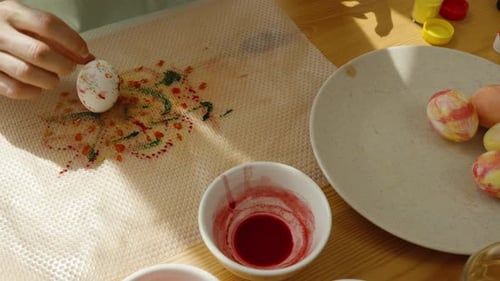 Hand rolling an Easter egg over a painted mat, with bowls of dye and white eggs on the table,