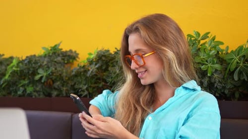 Happy Female Freelancer in Trendy Eyeglasses Using Smartphone at Cafe
