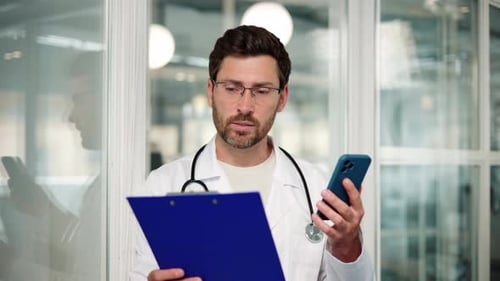 Male Doctor Using Smartphone and Holding Clipboard