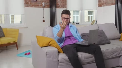Attractive Young Man Freelancer Sitting on Sofa Use Laptop Stay at Home Points His Finger at the