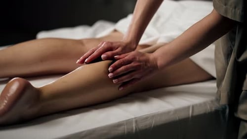 Close Up in a Massage Parlor on a Massage Table a Girl with Bare Legs Massage with Black Stones