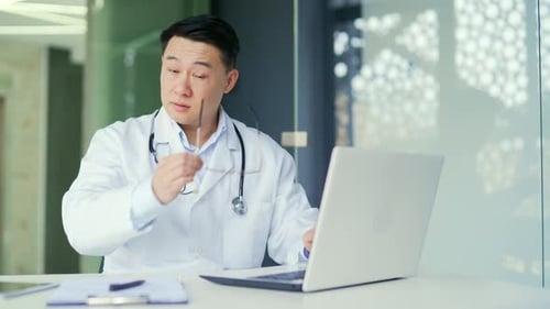 Overworked asian doctor suffering from eyestrain working on laptop while sitting in workplace