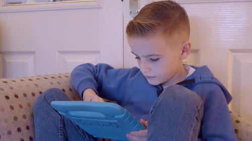 Close up of a child playing games on a tablet while sitting on a sofa at home.
