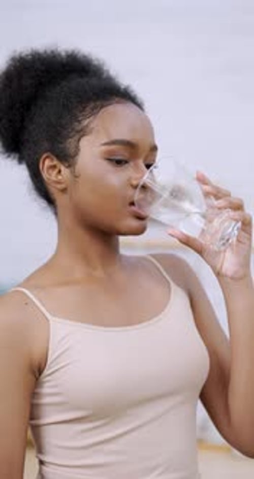 Adolescente africaine souriante et tenant un verre d'eau transparent dans une cuisine lumineuse...
