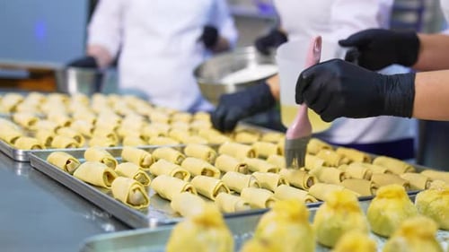 Unbaked croissants being prepared for baking in oven. Bakers cover the pastry with liquid. Close up.