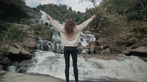 A Young Woman Raises Her Hands in the Air at a Majestic Waterfall in Northern Thailand