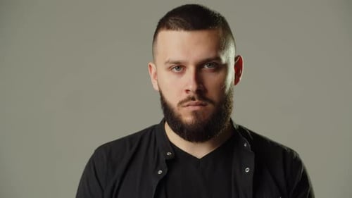 Portrait Shot of a Young Bearded Man Against a Gray Wall A Man in a Black Sweatshirt Poses for the