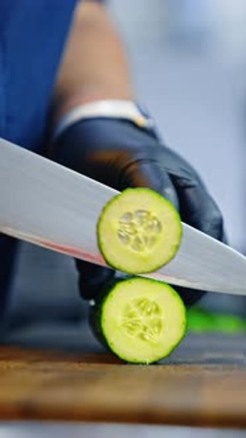 Unrecognized cook's hands in latex gloves cut the cucumber with a big sharp knife.