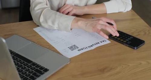 Junge Frau schreibt Steuererklärungsformular und schaut auf Laptop am Tisch im Wohnzimmer