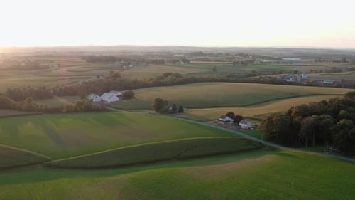 Aerial pullback reveals rolling hills, green farmland in countryside. Summer sunset on horizon.