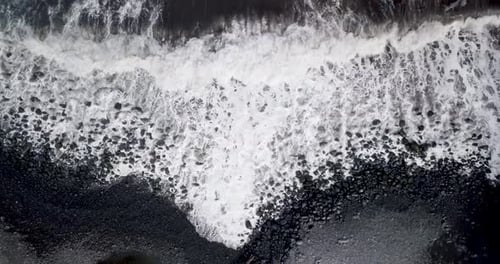 Cinematic top-down drone aerial of rhythmic waves crashing on the black volcanic sand of Maui beache