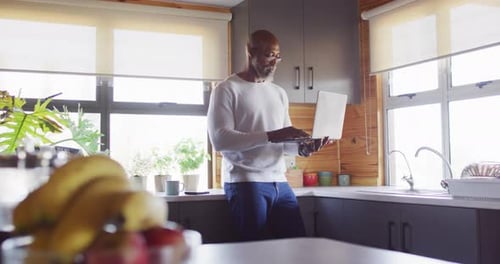 Happy senior african american man in log cabin, standing in kitchen and using laptop, slow motion