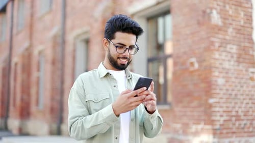 A handsome bearded student uses a smartphone while standing in the campus space near the university