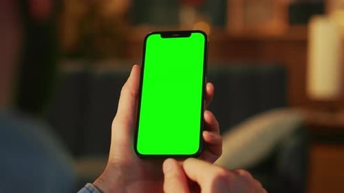 Männerhände halten Smartphone mit grünem Bildschirm auf verschwommenem Hintergrund des Wohnzimmers am Abend