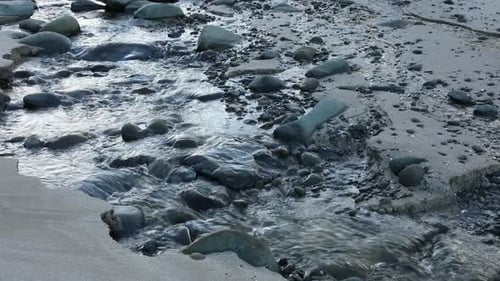 Inland river running into open sea water with seaweed next to sandy beach.