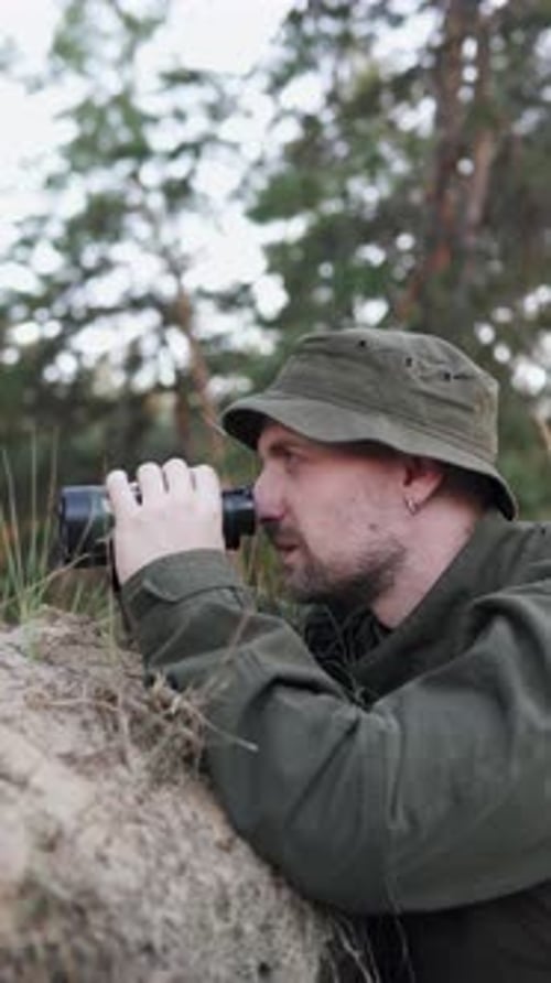 Soldier observing enemy through binoculars while hiding in forest position. Vertical video format.