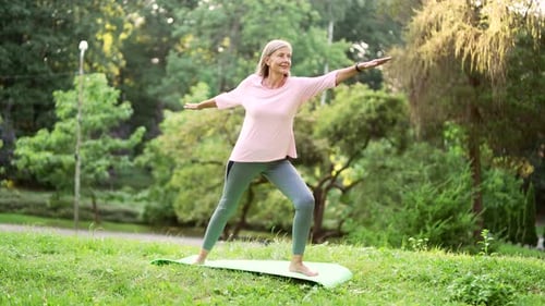 Senior active gray haired woman does gymnastics while standing on a mat in an urban city park.
