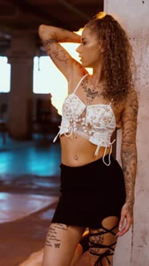 Vertical video of tattooed woman posing at warehouse pillar as fire ignites behind