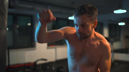 Alone Boxer is Working Out Blows on a Punching Bag in a Gym in Evening