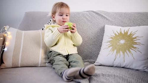 A Young Child Sits on a Cozy Sofa Enjoying a Green Apple with Curiosity and Contentment Surrounded
