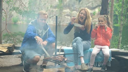 A Couple with a Child Grilling in the Forest on the Shore of the Lake