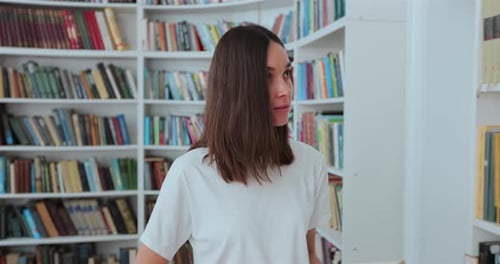Menina bonita caucasiana em camiseta branca caminha pelas prateleiras com livros na biblioteca