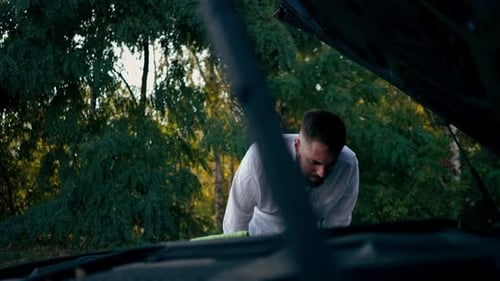 a young bearded man stands near a car with an open hood and tries to check a breakdown in the middle