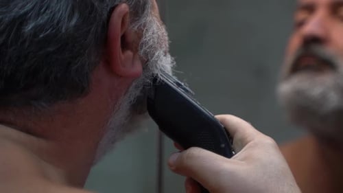 Man Trimming Beard with Electric Razor in Mirror