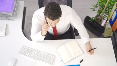 Businessman talking on phone in office and taking notes in notebook.