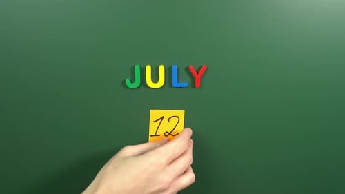 Hand sticking a sticker 12 July calendar day on school board. 12 date of July. Twelfth day July