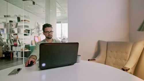 Glücklicher junger Mann mit Brille und Lächeln, während er an seinem Laptop arbeitet, um all seine Geschäfte zu erledigen