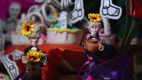 altar de los muertos con un par de catrinas en primer plano sosteniendo una flor amarilla y una vasija de barro,