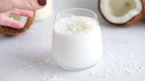 A Hand Picks Up a Glass of Fresh Coconut Milk Next to a Coconut Shell Cut in Half